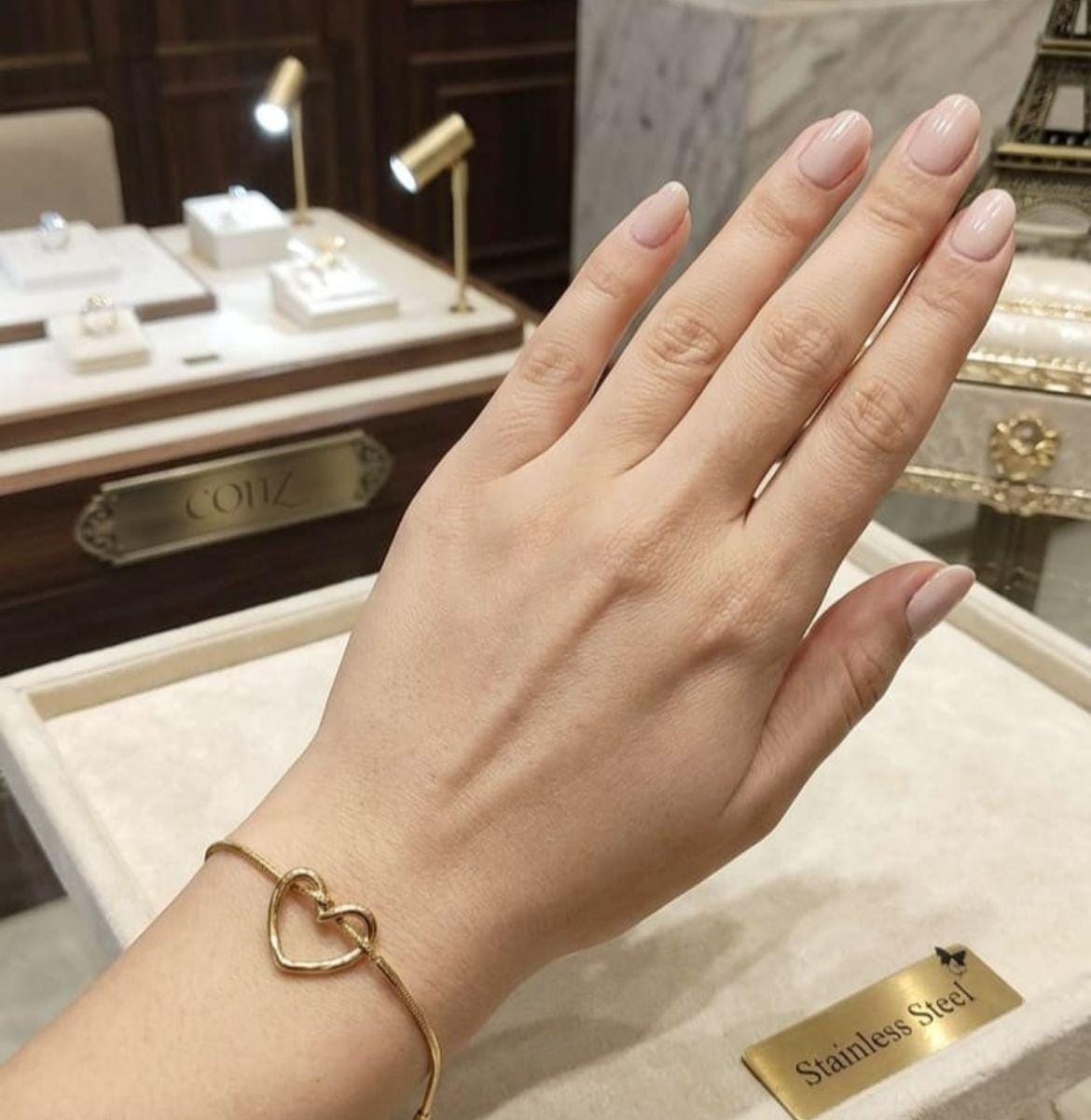 A gold heart-link bracelet worn on a woman's wrist, shown in a jewelry store setting with display cases and warm lighting in the background.