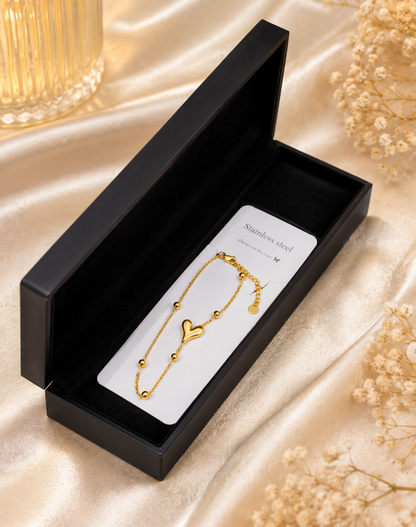 A delicate gold bracelet with a heart charm and ball beads, presented in an open black gift box on a white card labeled 'Stainless steel'. The box is set on a beige satin fabric background with dried flowers.