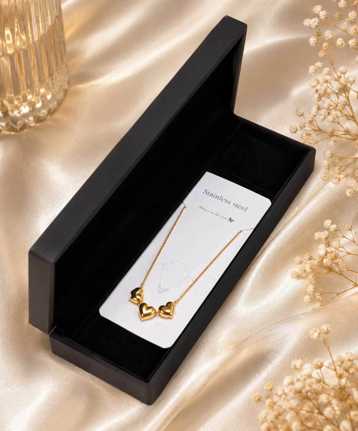 A gold necklace with three small heart charms displayed on a white card inside an open black presentation box, set on a champagne satin fabric with dried floral decor.
