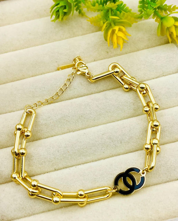 Gold bracelet with a black clasp on a light wooden surface with green leaves.