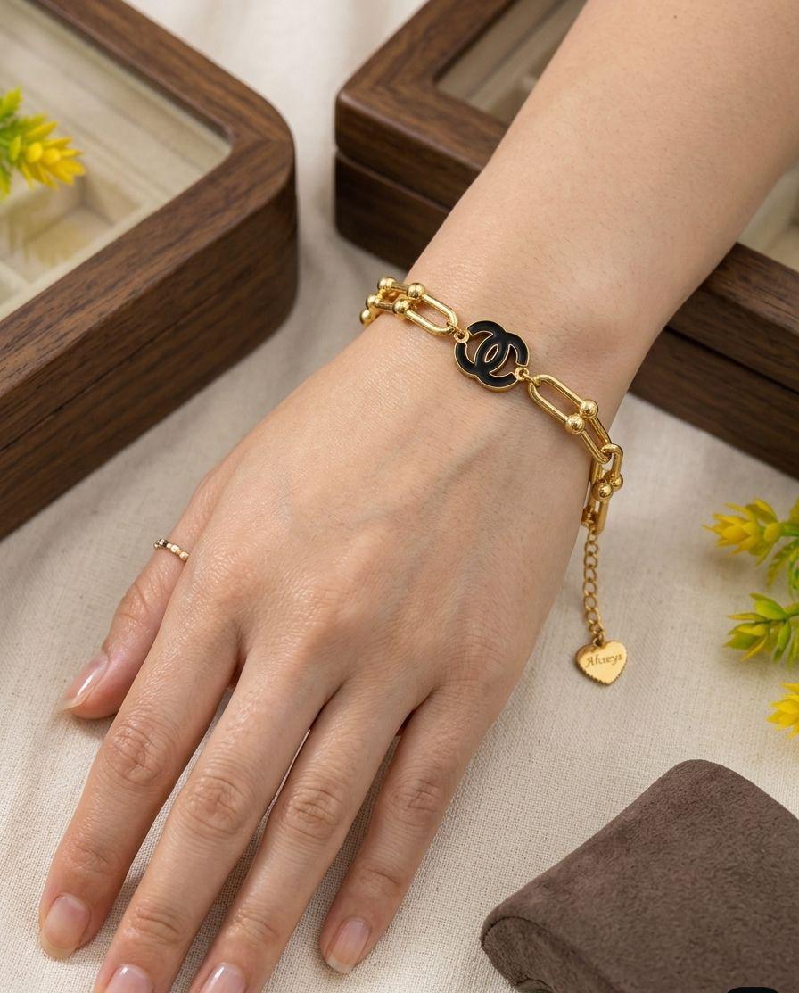 Gold bracelet with a heart charm on a wrist, with a wooden box and yellow flowers in the background.