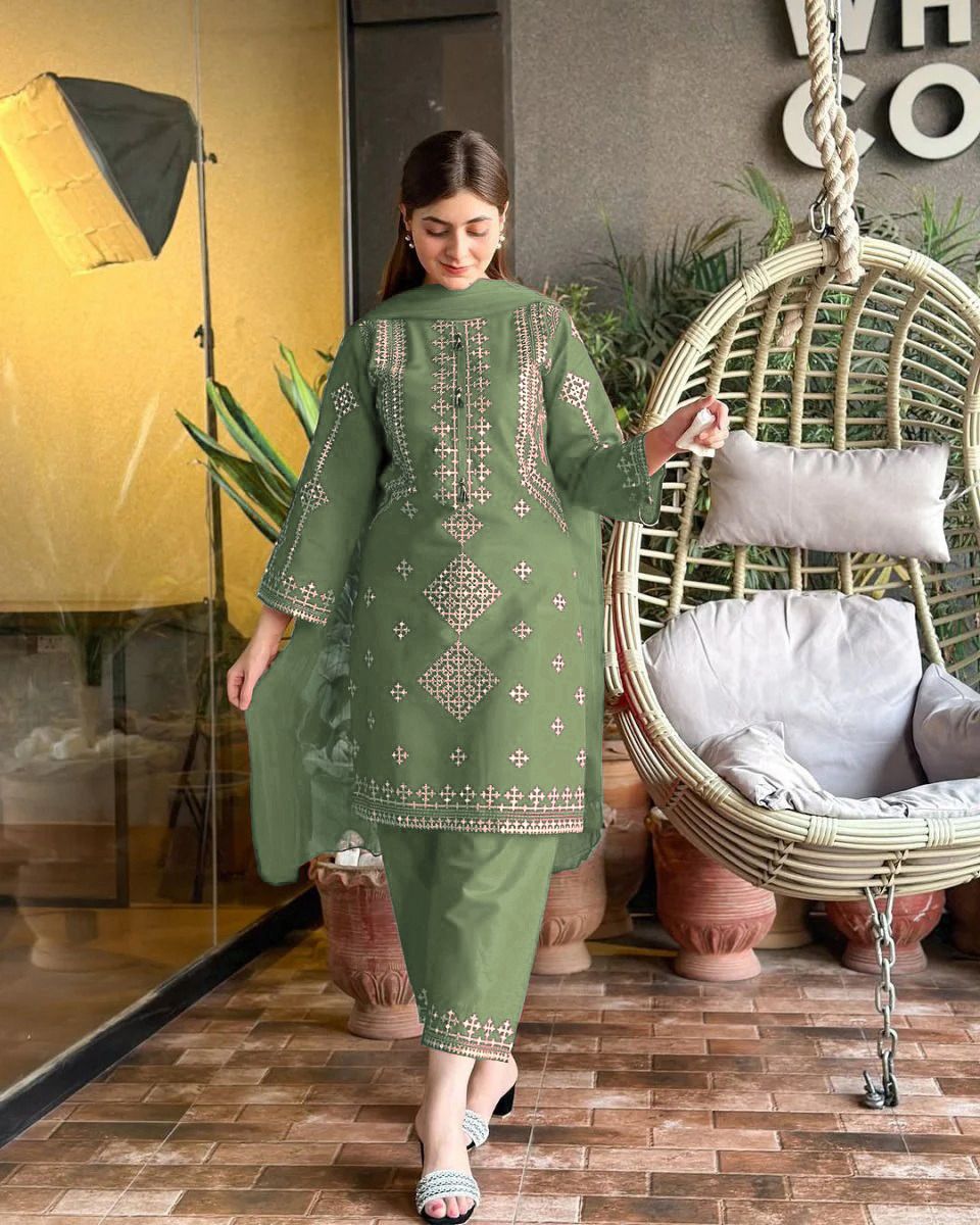 Woman in a green embroidered dress standing in a room with a wicker chair and plants.
