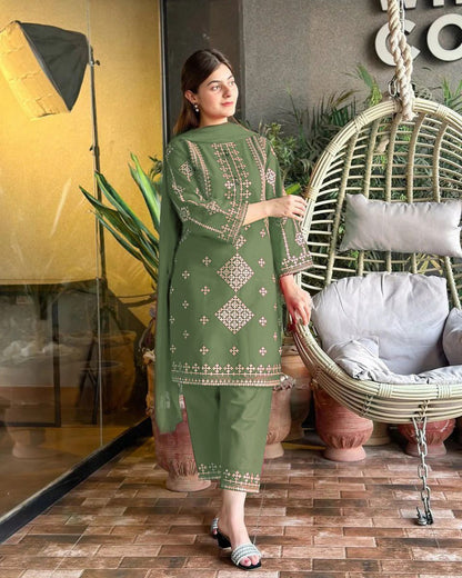 Woman in a green embroidered outfit standing next to a wicker chair indoors.
