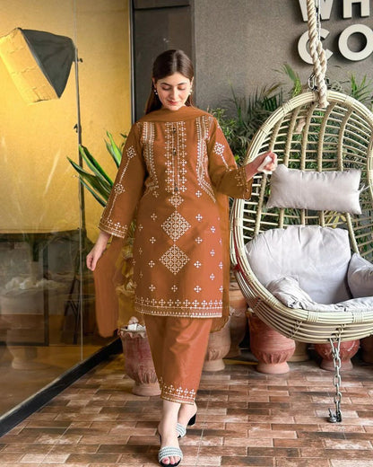 Woman in a brown embroidered outfit standing in a room with a wicker chair and plants.