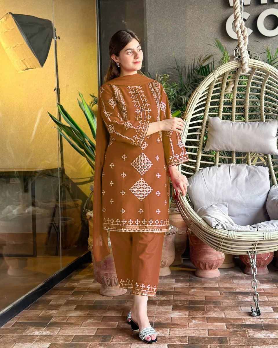 Woman in a brown embroidered outfit standing next to a wicker chair with cushions.