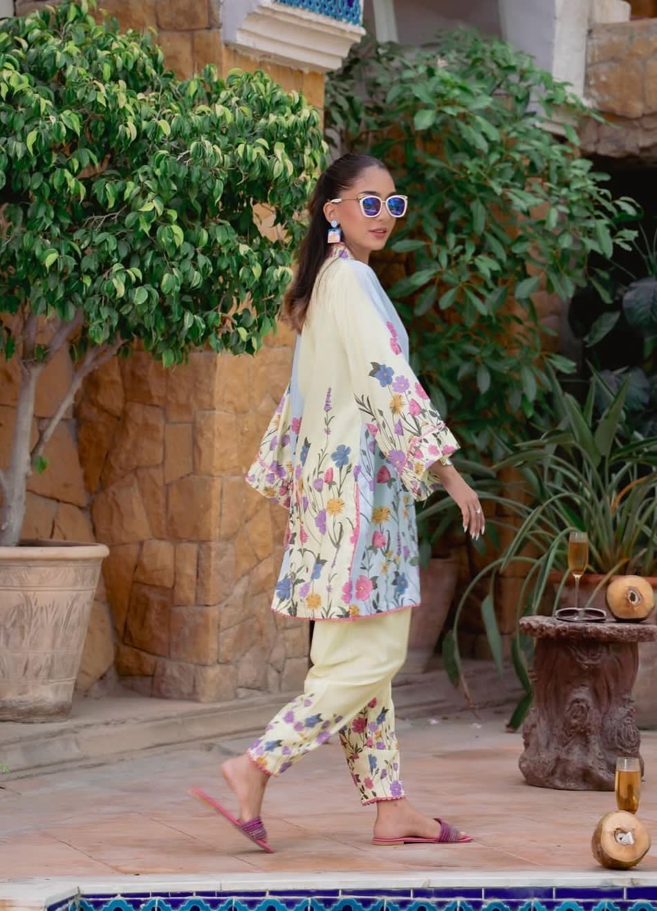 Woman in floral outfit walking outdoors with plants and stone wall in background