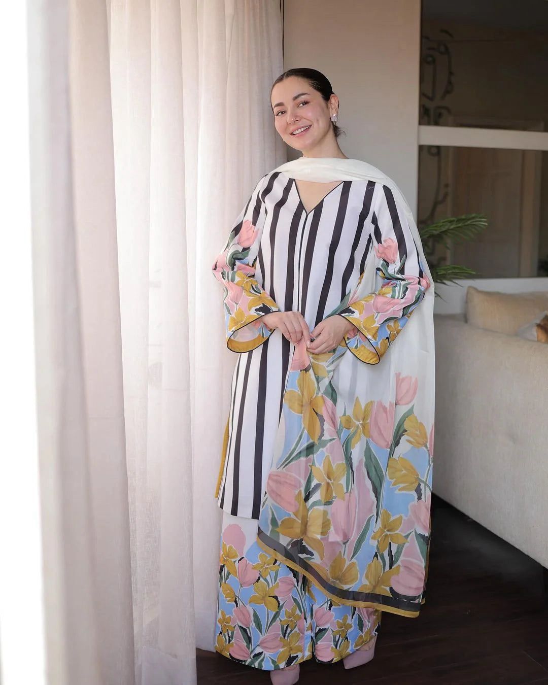 Woman wearing a floral and striped dress in a room with curtains and a couch.