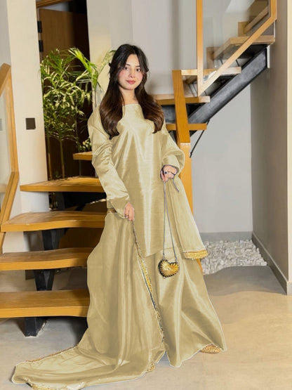 Woman in a long, light-colored dress standing indoors with wooden stairs and plants in the background.