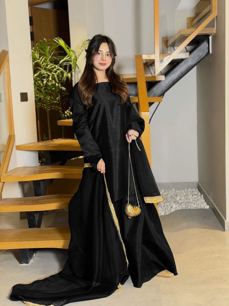 Woman in a black traditional outfit with gold trim standing in a home setting.