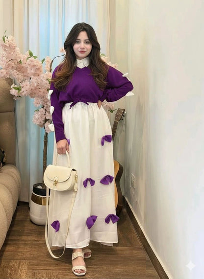 Woman in a purple sweater and white skirt with purple accents standing in a room with flowers and furniture.