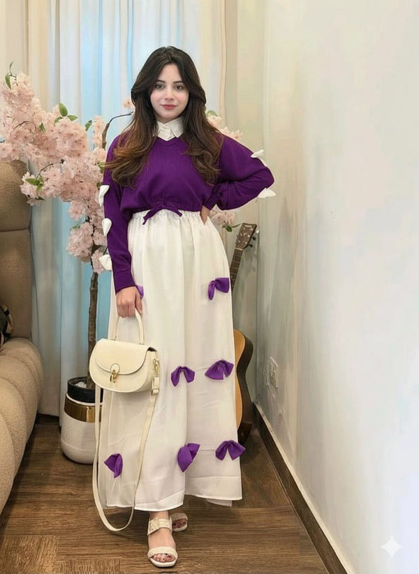 Woman in a purple sweater and white skirt with purple accents standing in a room with flowers and furniture.