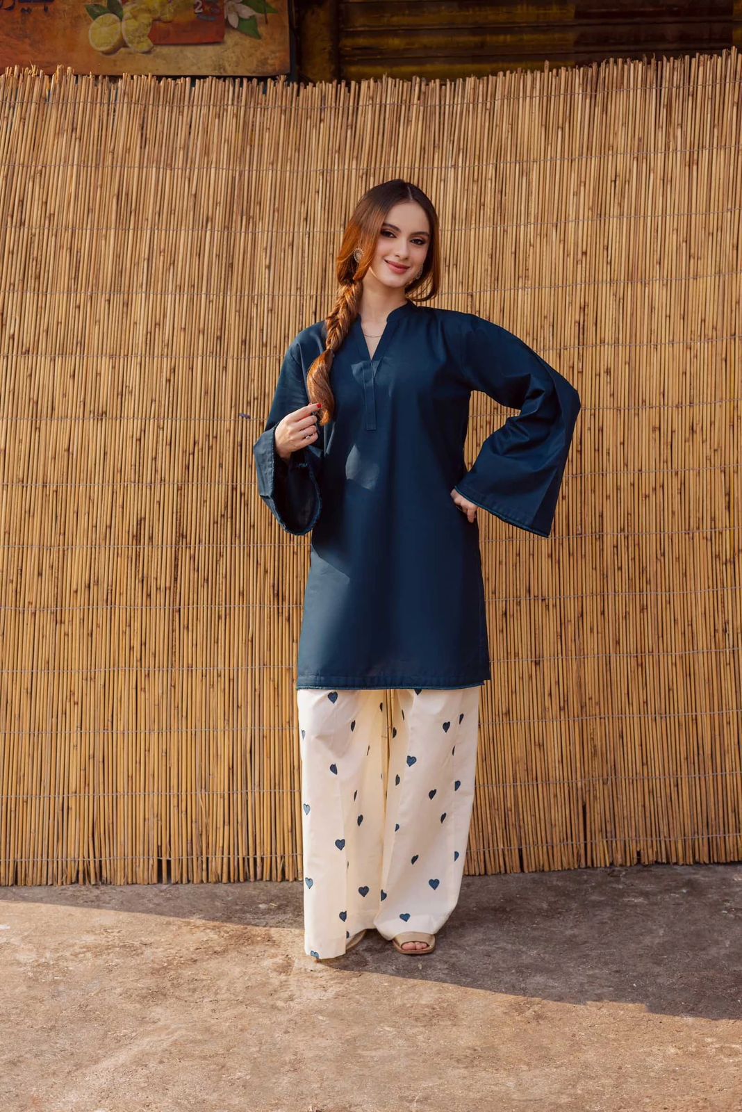 Woman wearing a dark blue top and light-colored pants with heart patterns against a bamboo wall.