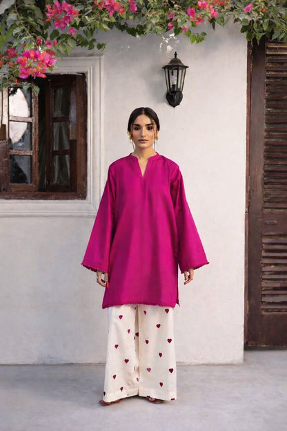 Woman wearing a bright pink top and white pants with heart patterns, standing in front of a white wall with flowers.