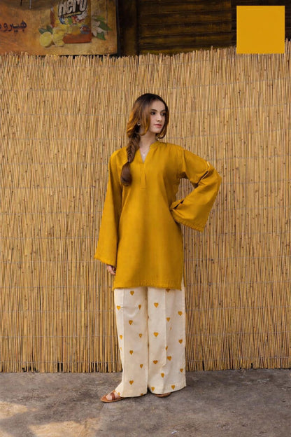 Woman wearing a yellow top and white pants with a floral pattern against a bamboo wall.