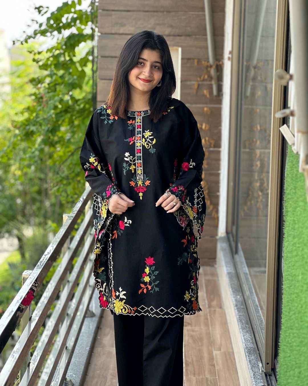 Woman wearing a black embroidered outfit standing on a balcony with greenery in the background