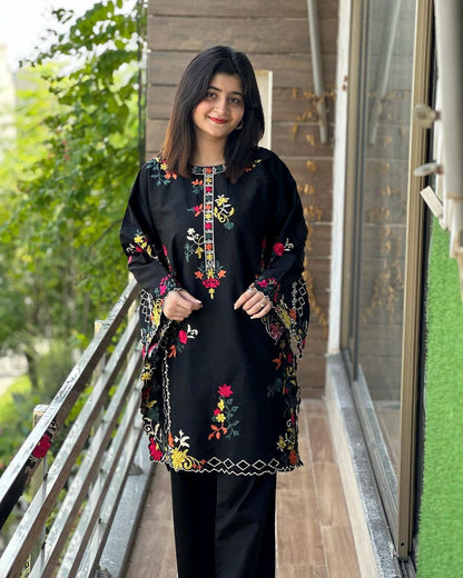 Woman wearing a black embroidered outfit standing on a balcony with greenery in the background
