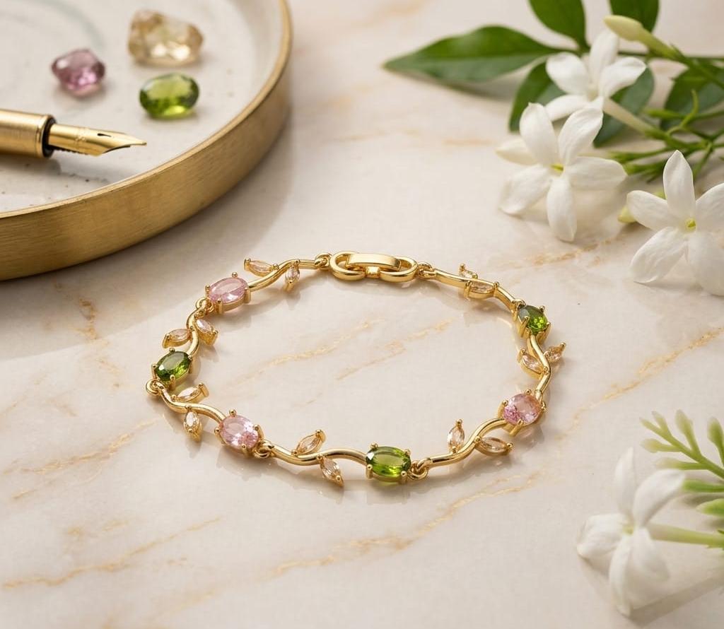 Gold bracelet with pink and green gemstones on a marble surface with flowers and a pen.
