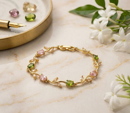 Gold bracelet with pink and green gemstones on a marble surface with flowers and a pen.