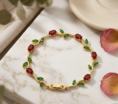 Gold bracelet with red and green gemstones on a marble surface with pink petals.