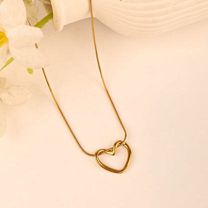 A gold heart-shaped pendant necklace with a delicate chain, displayed on a soft cream background with a white textured vase and white flowers as props.