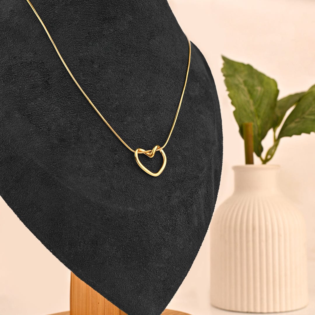 Gold necklace with a heart-shaped pendant on a black stand with a beige background and plant.