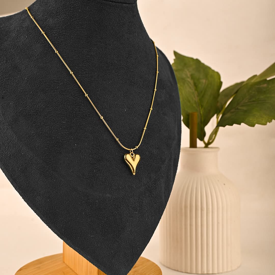 Gold necklace with a heart pendant on a black mannequin against a neutral background with a vase and plant.