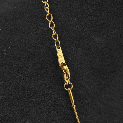 Close-up of a gold-tone chain necklace with a lobster clasp and tag, photographed against a dark textured background. The tag is engraved with 'GUESS'.