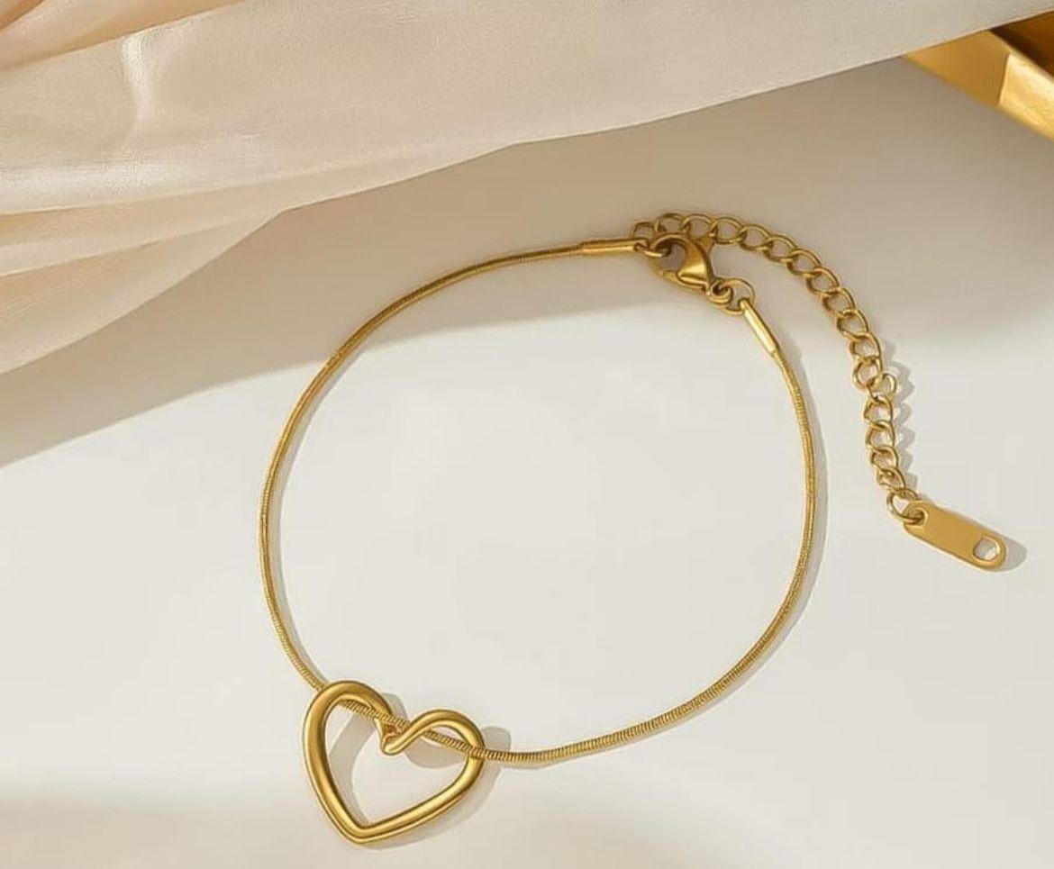 A gold chain bracelet with a double-heart pendant, displayed on a clean white surface with soft beige fabric in the background.