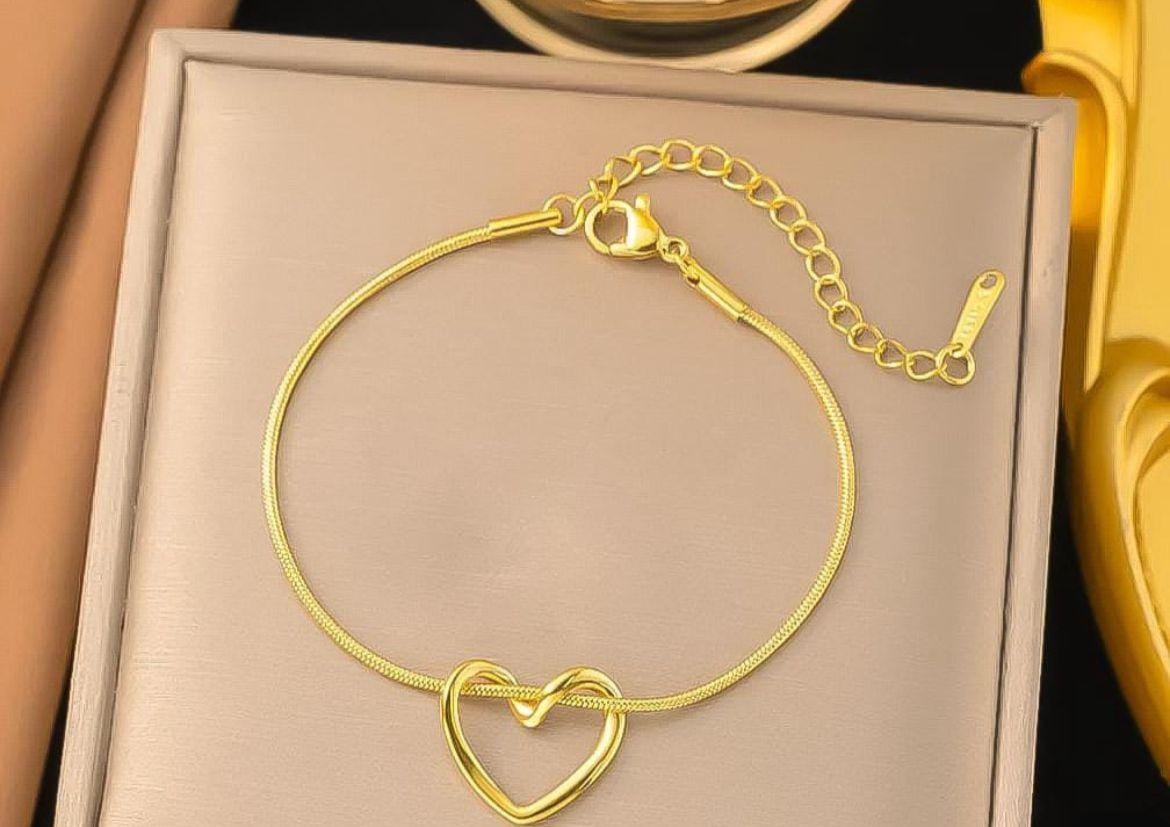 A gold chain bracelet with a heart-shaped infinity link pendant, displayed on a beige jewelry box.