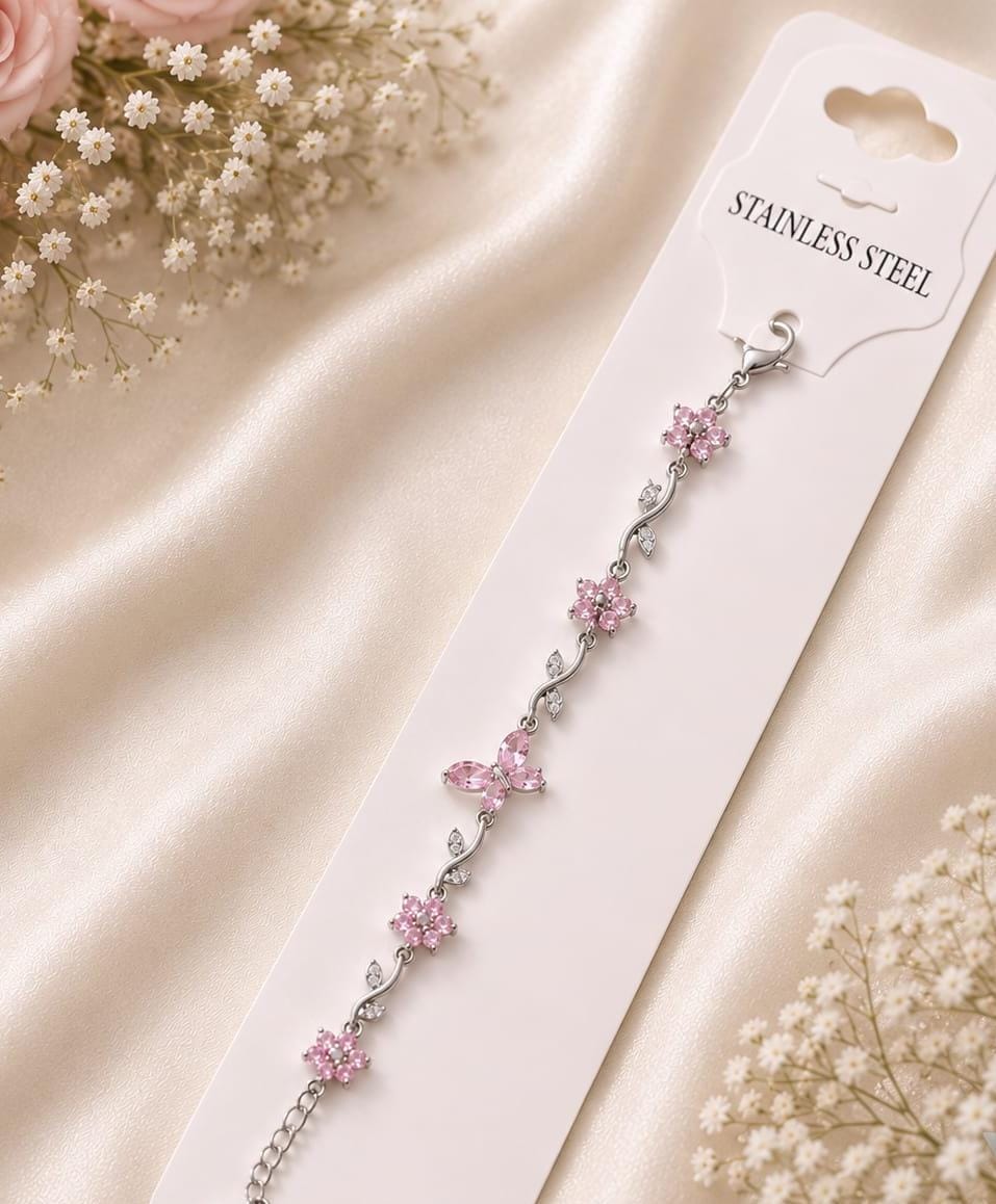 A silver-toned stainless steel chain bracelet with pink flower and teardrop crystal embellishments, displayed on a white card labeled 'STAINLESS STEEL' against a soft cream satin background with small white flowers.