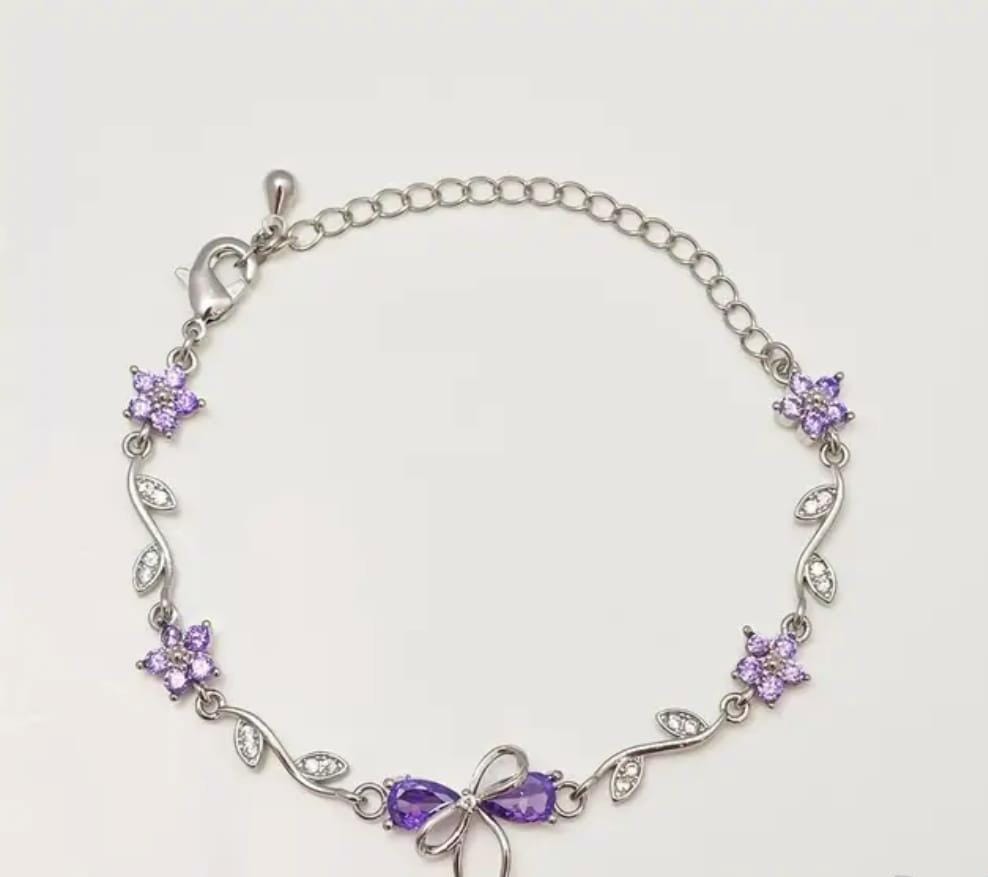 A silver chain bracelet featuring purple crystal flowers and a bow at the center, displayed on a light off-white background.