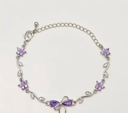 A silver chain bracelet featuring purple crystal flowers and a bow at the center, displayed on a light off-white background.
