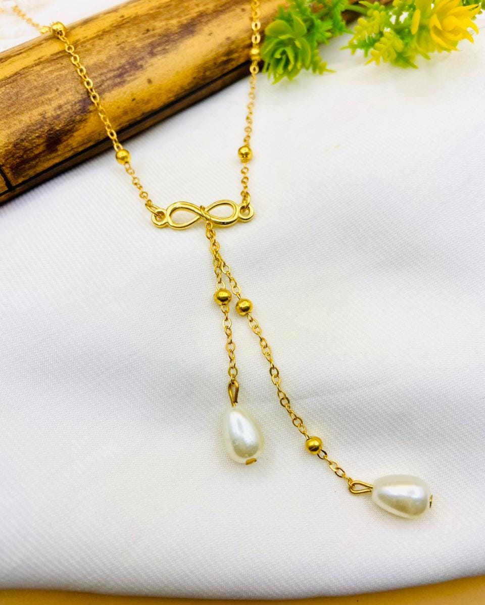 A gold-toned lariat-style necklace with an infinity symbol at the center, small bead accents, and two dangling pearl-like teardrop charms, displayed on a white fabric surface with a wooden prop and green flowers.