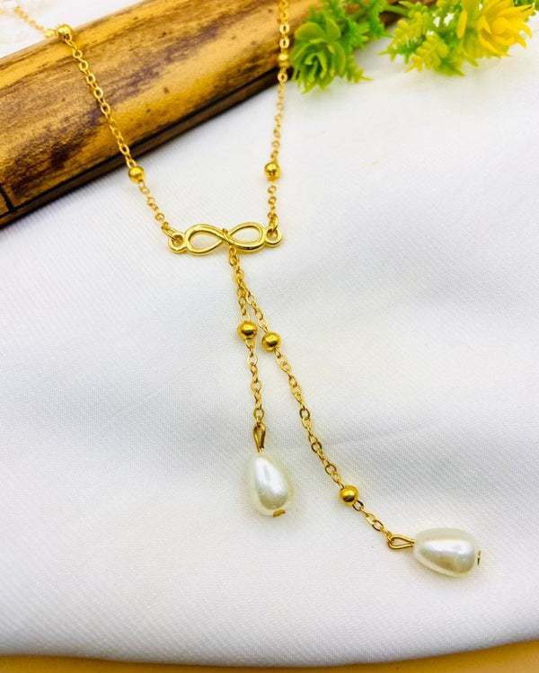 A gold-toned lariat-style necklace with an infinity symbol at the center, small bead accents, and two dangling pearl-like teardrop charms, displayed on a white fabric surface with a wooden prop and green flowers.