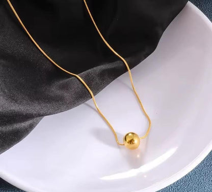 A delicate gold chain necklace with a small, polished gold sphere pendant, draped over a white plate with a black satin cloth underneath.