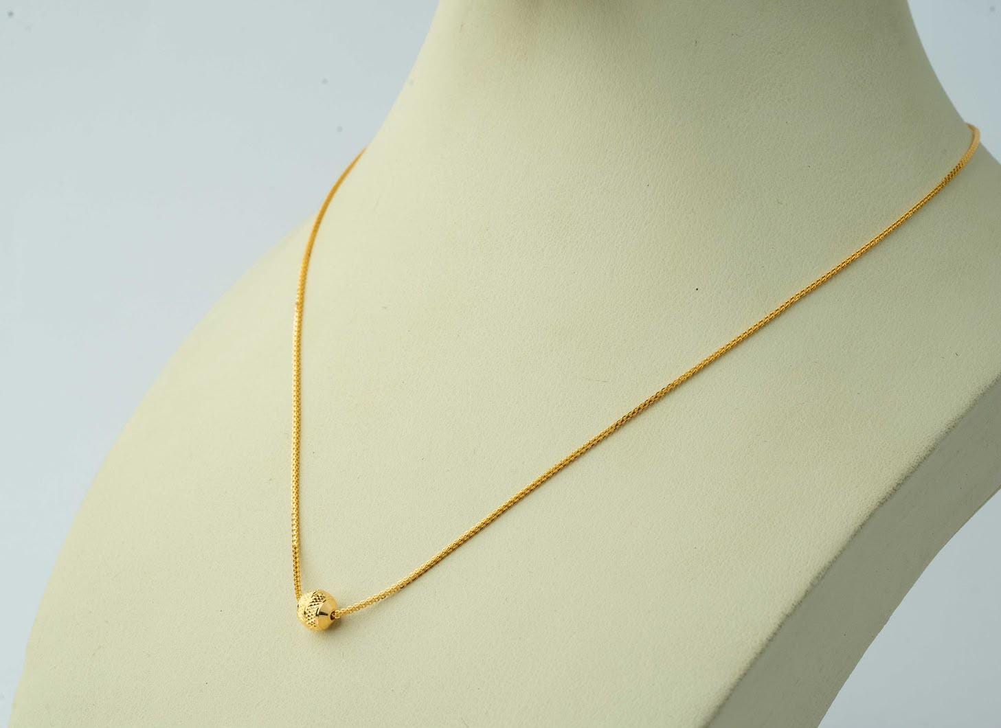 A delicate gold chain necklace with a small, textured gold bead pendant, displayed on a cream-colored mannequin bust against a light gray background.
