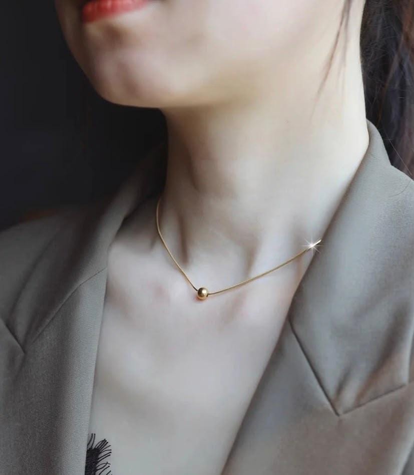 A close-up lifestyle shot of a model wearing a delicate gold chain necklace with a small spherical bead pendant, styled with a taupe blazer.
