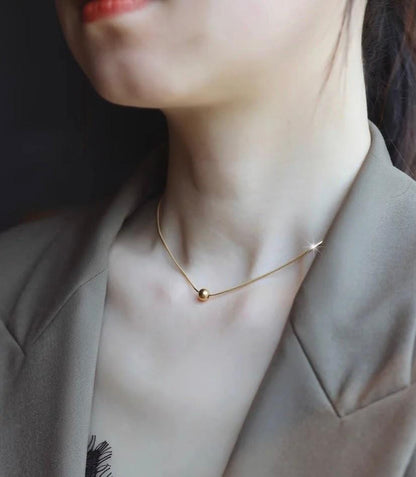 A close-up lifestyle shot of a model wearing a delicate gold chain necklace with a small spherical bead pendant, styled with a taupe blazer.