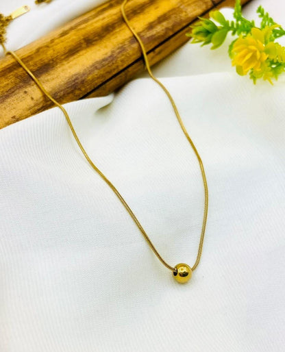 A delicate gold snake chain necklace with a small, polished gold bead pendant, displayed on a textured white fabric with a wooden prop and yellow flowers in the background.