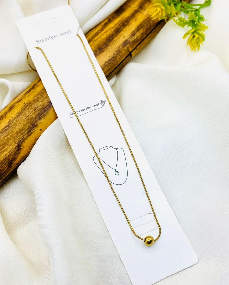 A stainless steel chain necklace with a small gold ball pendant, displayed on its white retail card packaging. The card is laid on a white fabric background with a decorative wooden prop and small yellow flowers.