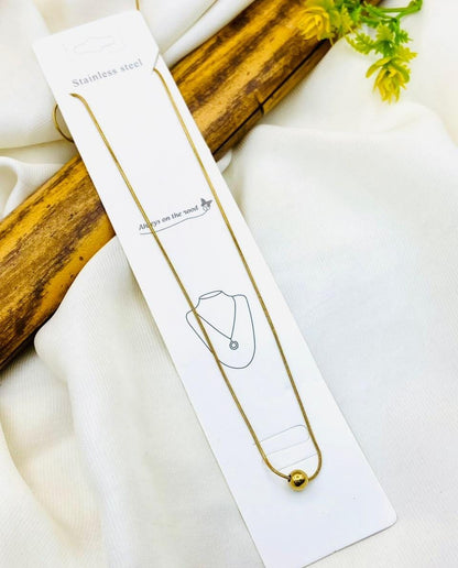 A stainless steel chain necklace with a small gold ball pendant, displayed on its white retail card packaging. The card is laid on a white fabric background with a decorative wooden prop and small yellow flowers.