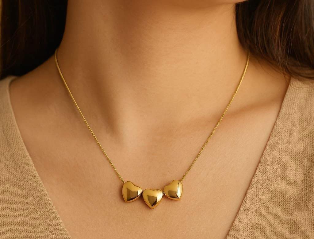 A close-up of a model wearing a delicate gold chain necklace with three small, polished gold heart charms.
