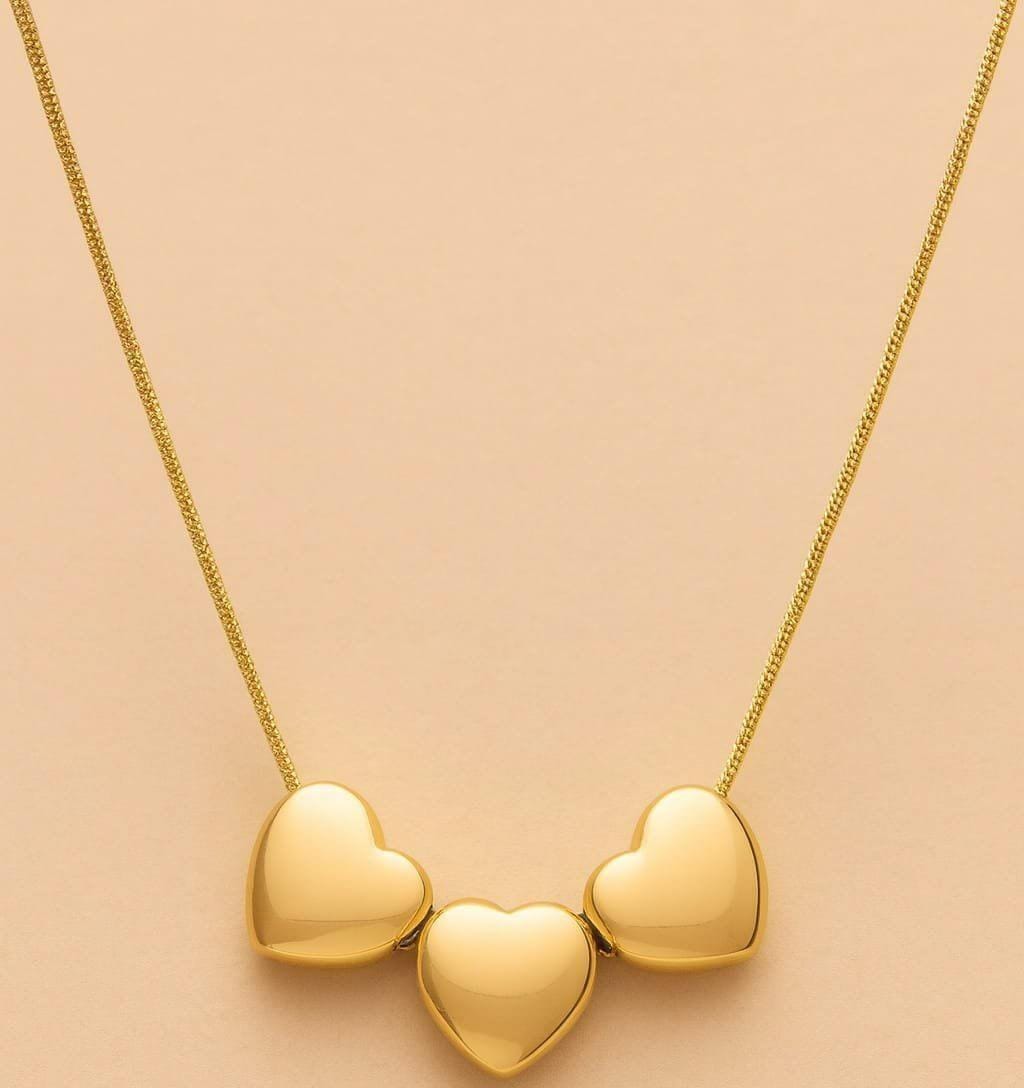 A gold necklace with three polished heart-shaped pendants on a delicate chain, displayed against a soft peach-pink background.