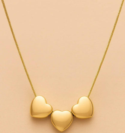 A gold necklace with three polished heart-shaped pendants on a delicate chain, displayed against a soft peach-pink background.