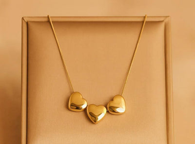 A gold necklace with three small, polished heart-shaped pendants displayed on a textured gold jewelry box.