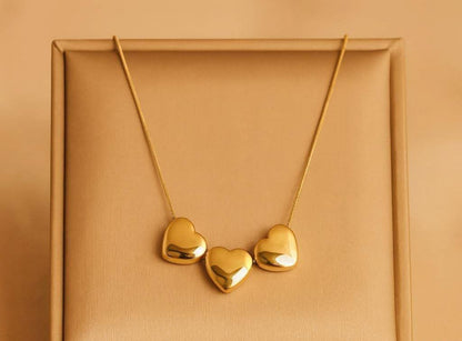 A gold necklace with three small, polished heart-shaped pendants displayed on a textured gold jewelry box.