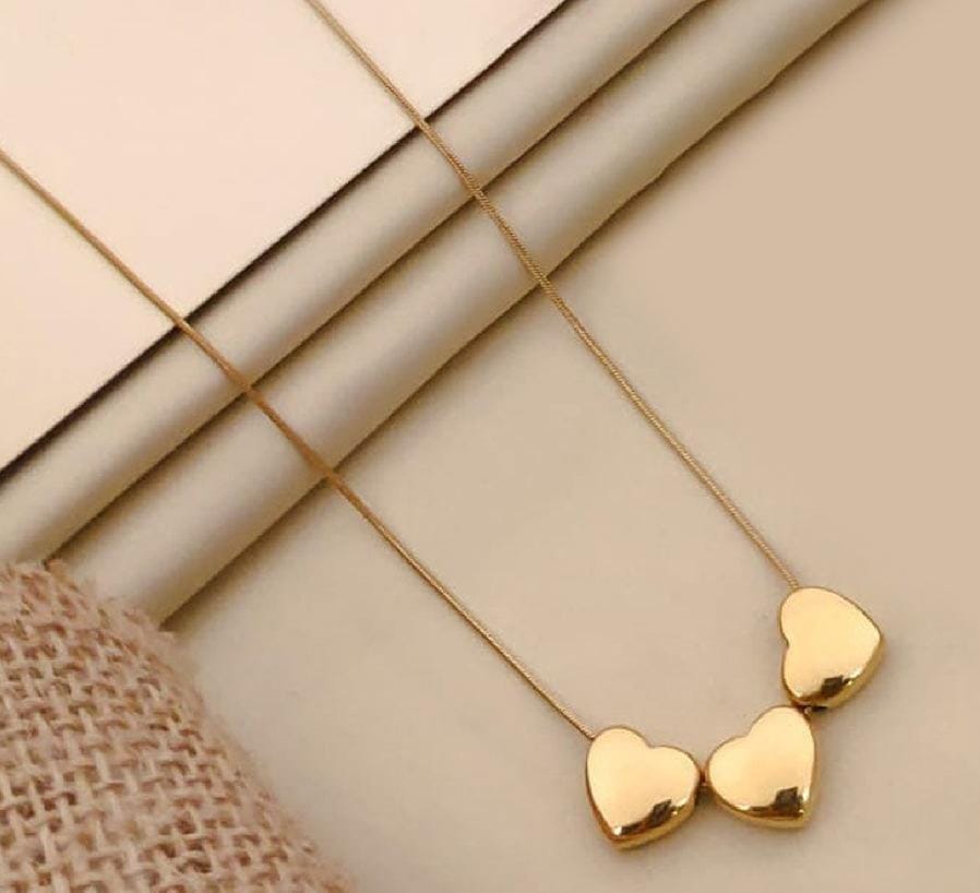A gold heart pendant necklace with three small, polished heart charms on a delicate chain, displayed on a neutral beige background with a textured burlap fabric and folded paper.