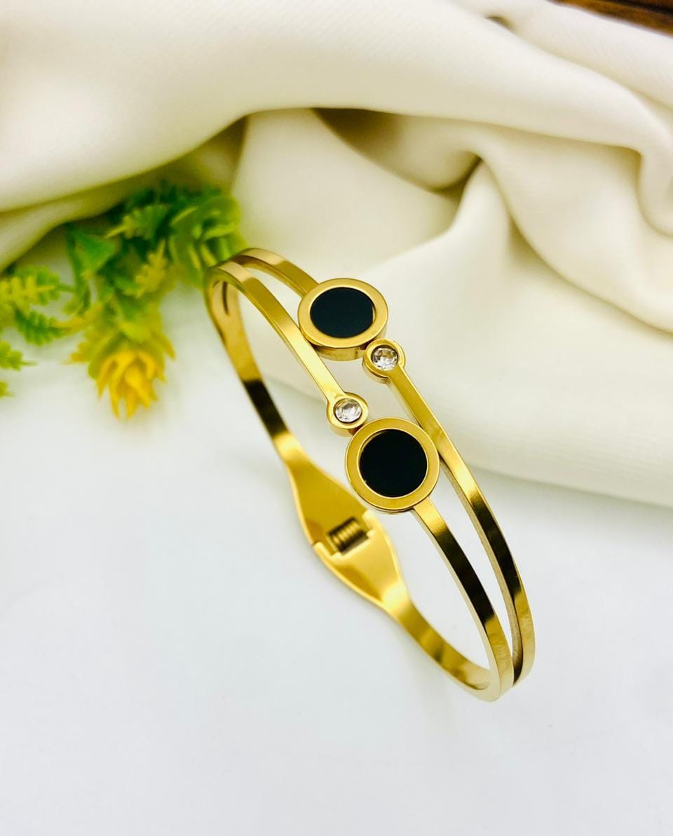 A gold-toned double-band cuff bracelet with two black circular inlays and a small clear rhinestone accent, displayed on a white surface with draped cream fabric and greenery in the background.