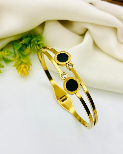 A gold-toned double-band cuff bracelet with two black circular inlays and a small clear rhinestone accent, displayed on a white surface with draped cream fabric and greenery in the background.