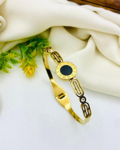 A gold-plated bangle bracelet with a central round black inlay and decorative chain-like links, styled on a white surface with draped cream fabric and small green leaves.